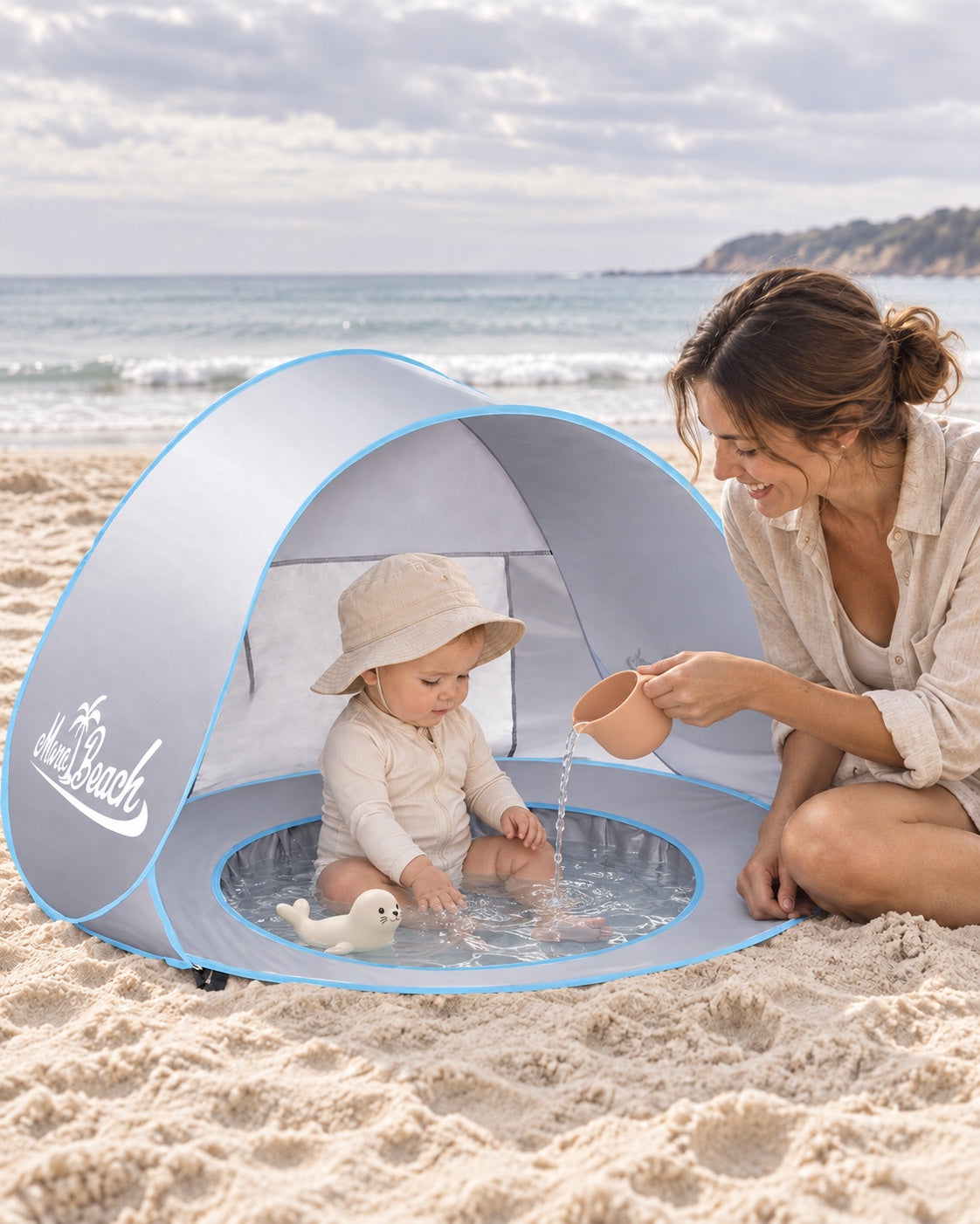 A mother beside her baby playing in the shallow water pool of a silver MonoBeach beach tent
